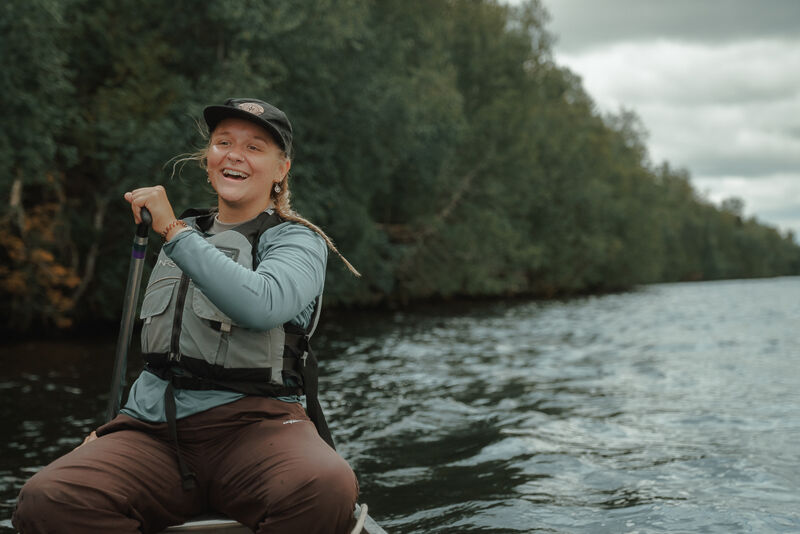 A woman wearing a life vest and a cap is paddling a canoe on a lake. She is smiling and looking to the side. The background shows a lush green forest and a cloudy sky. The water is calm with small ripples. She seems to be enjoying a peaceful day on the water.